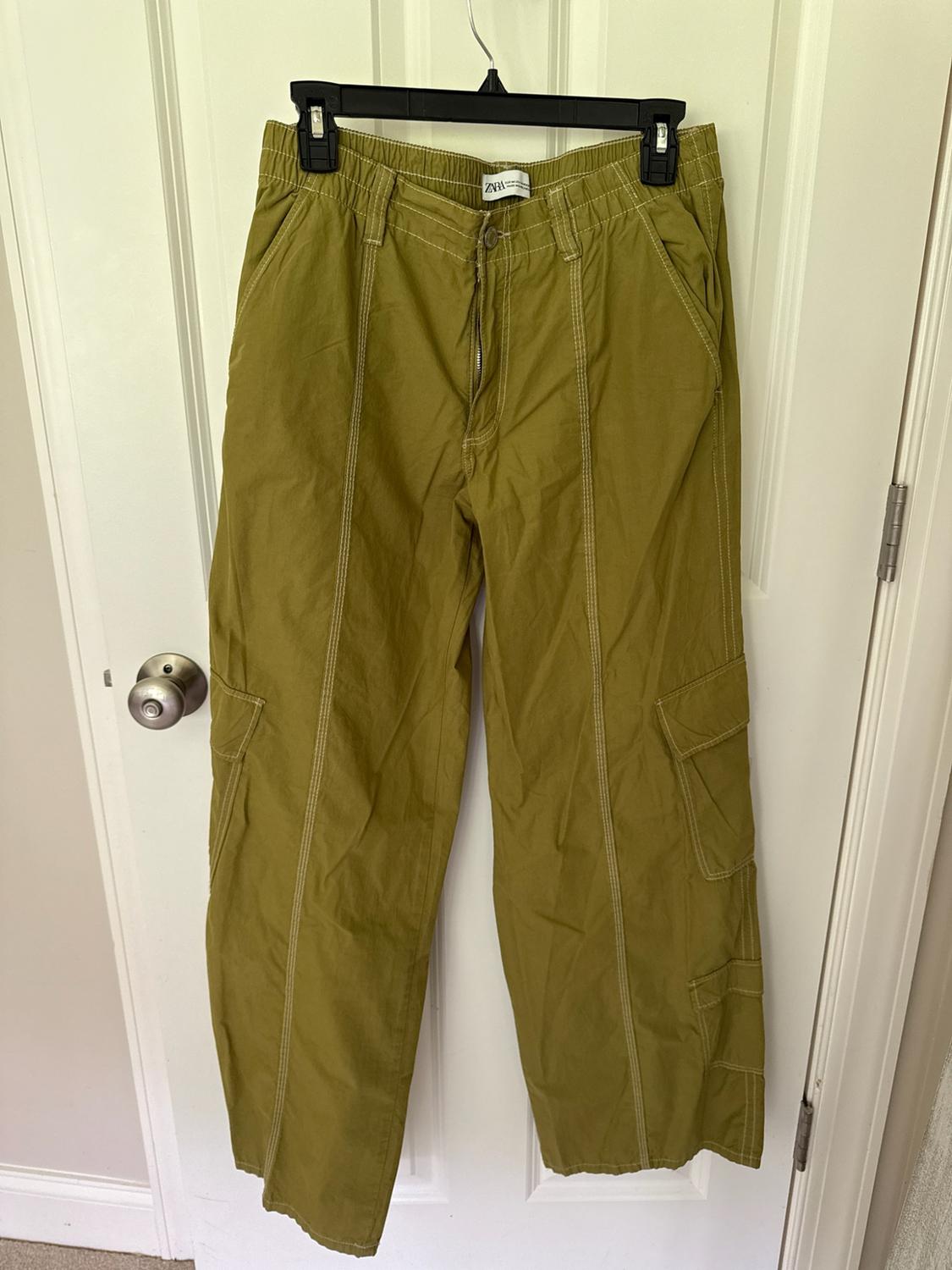 Olive green cargo pants with white stitching, hanging on a hanger over a white door. Text: ZARA.