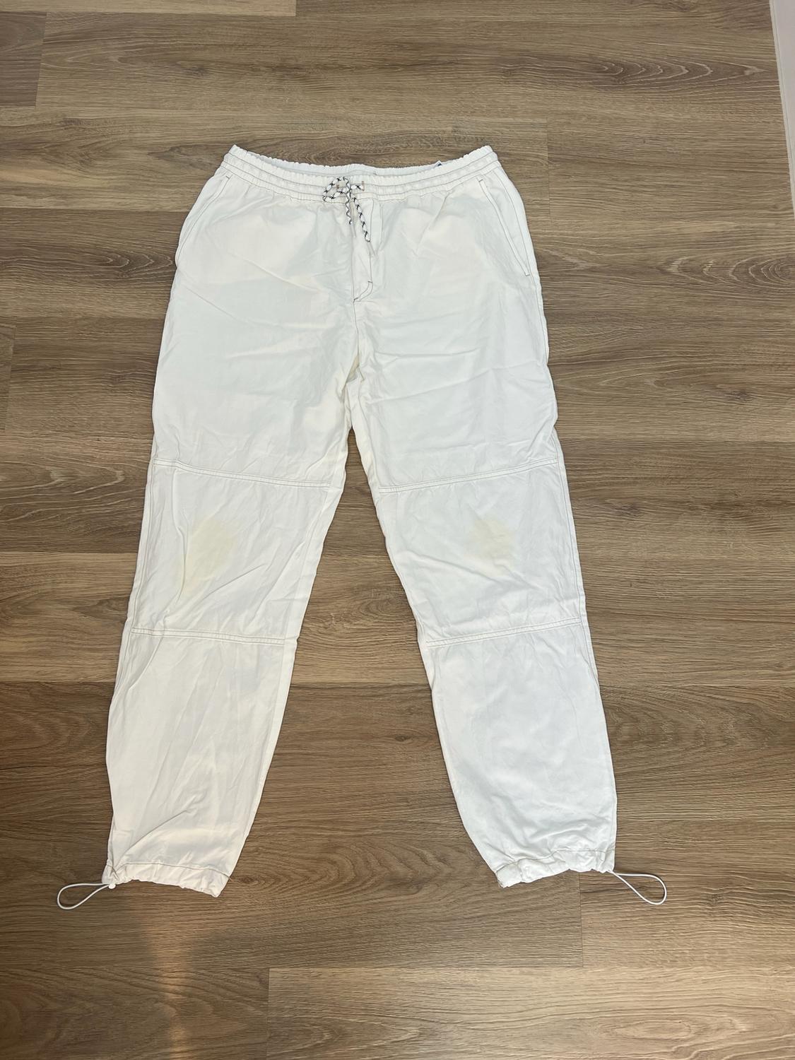 White trousers with an elasticated waist, adjustable drawstrings, knee seams and gathered hems, on a wooden floor.