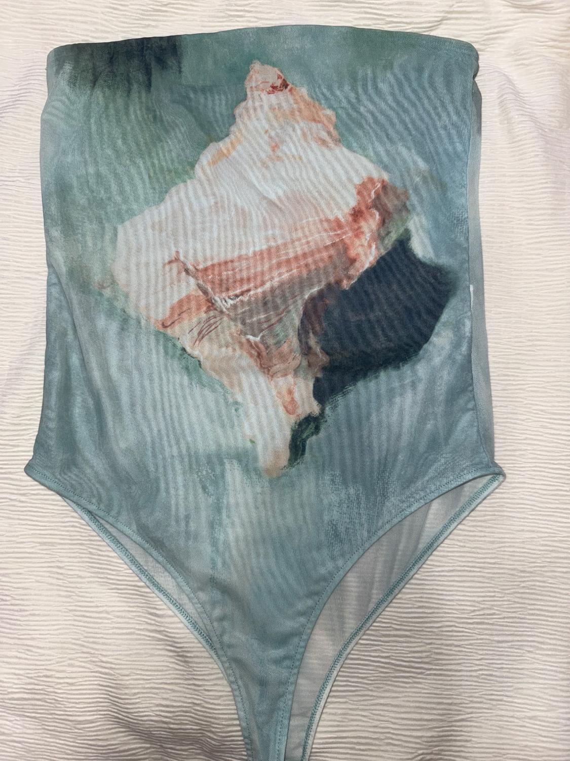 Light blue bodysuit with an abstract print in earthy and green tones, on a textured white background.