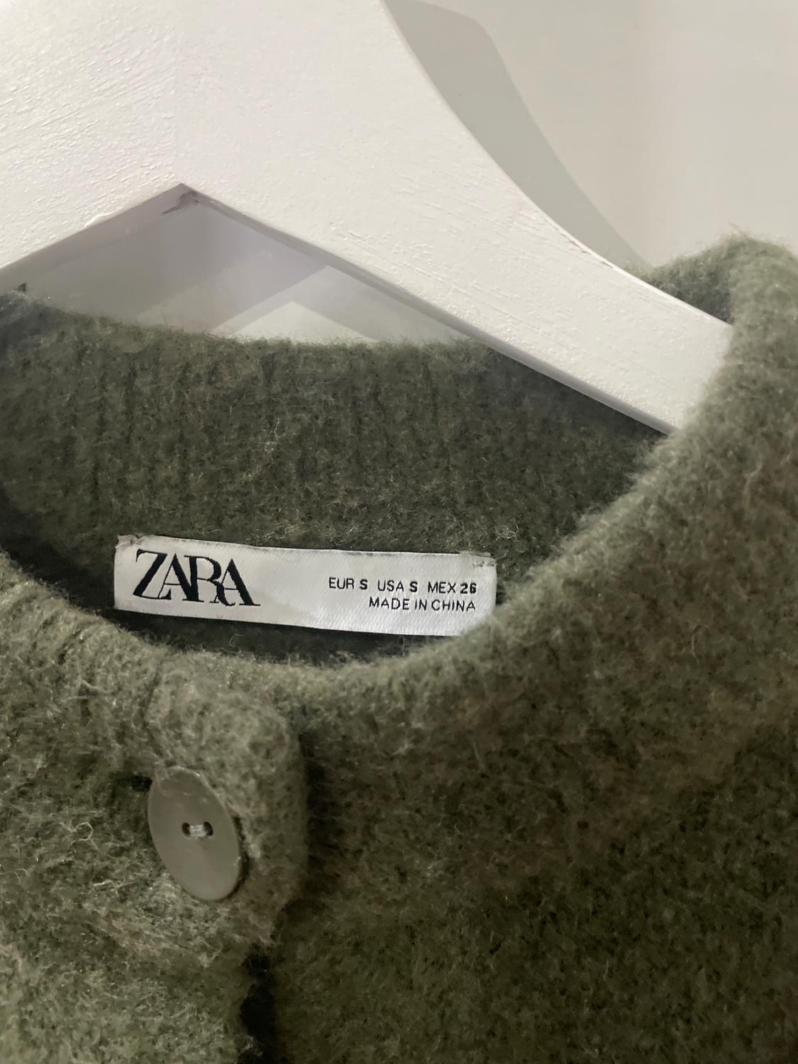 Label from a green wool garment with a button, hanging on a white hanger. Visible text: "ZARA EUR S USA S MEX 26 MADE IN CHINA".