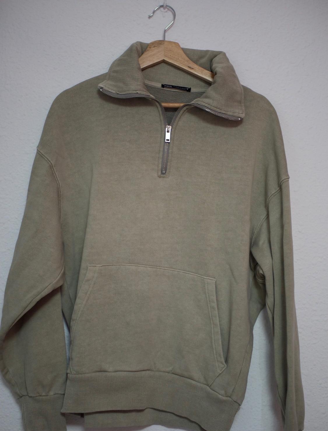 Khaki green long-sleeved half-zip sweatshirt with a ribbed high neck and kangaroo pocket, hanging on a hanger against a white background.