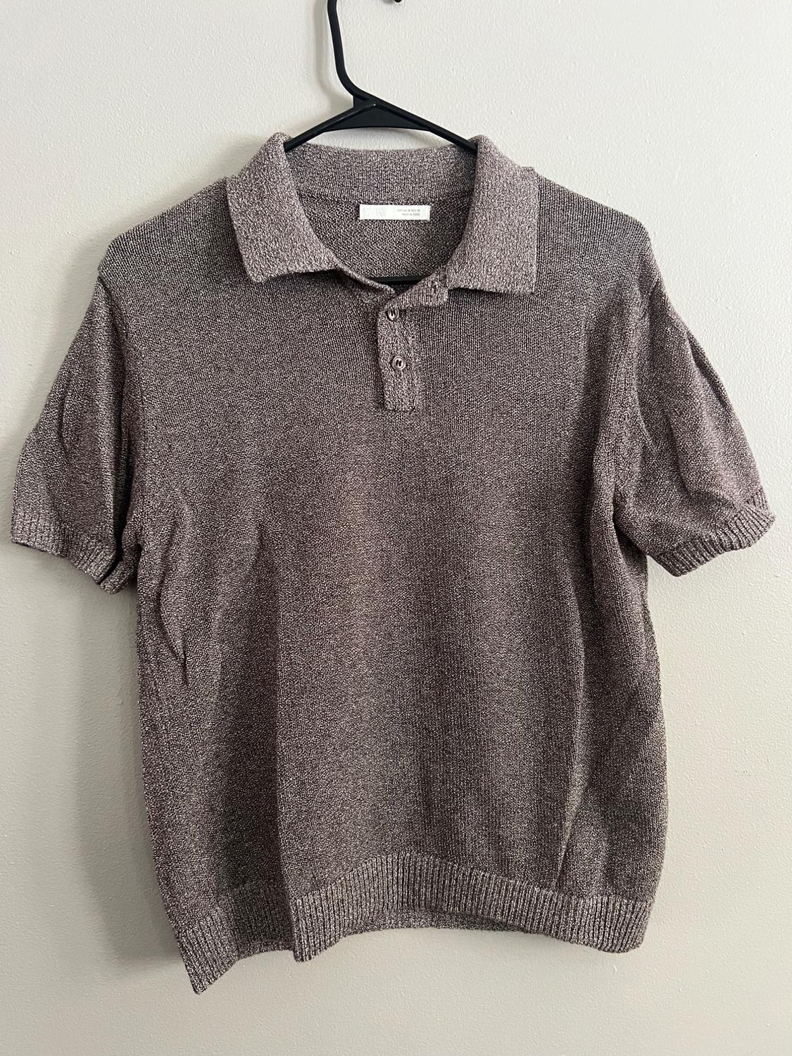 Gray short-sleeved polo shirt hanging on a black hanger against a white wall. Knit fabric with two buttons at the neck.