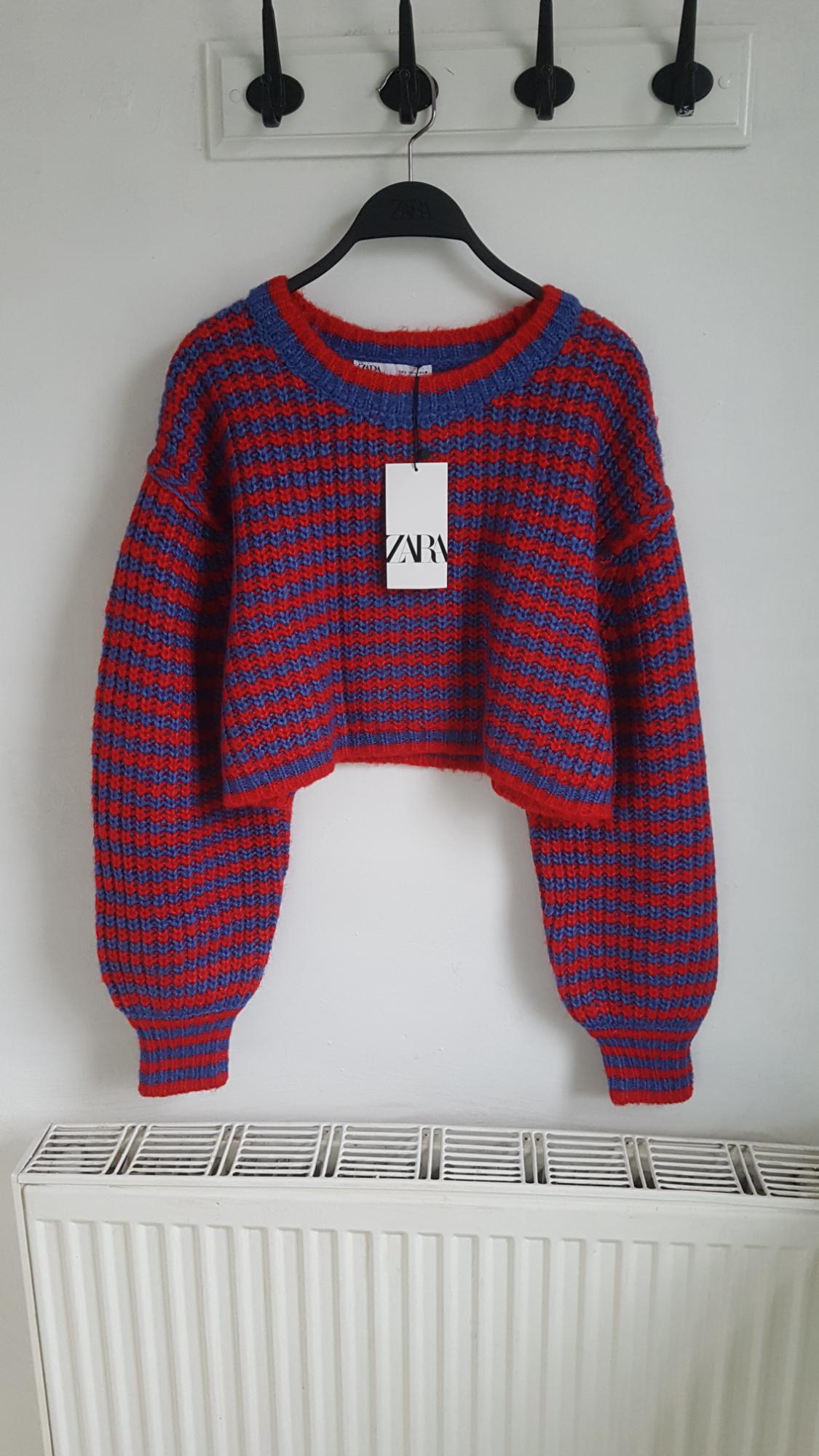 Blue and red knitted cropped jumper with Zara label, hanging on a black hanger against a white wall.