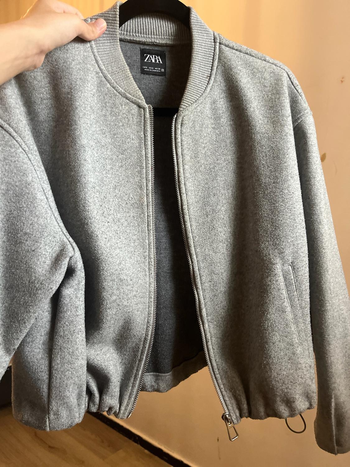 Hand holding a grey bomber jacket with a zip on a black hanger. Inner label with the text ZARA. Light background.