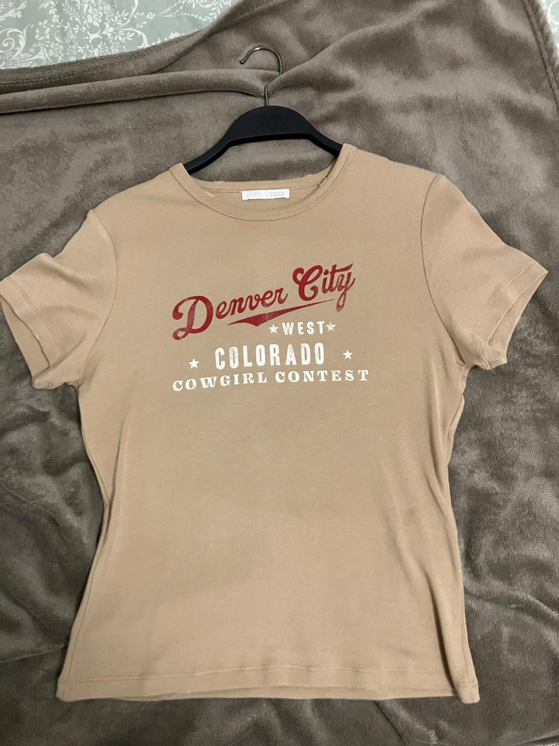 Beige short-sleeved t-shirt on a blanket. Text: Denver City * WEST * COLORADO * COWGIRL CONTEST.