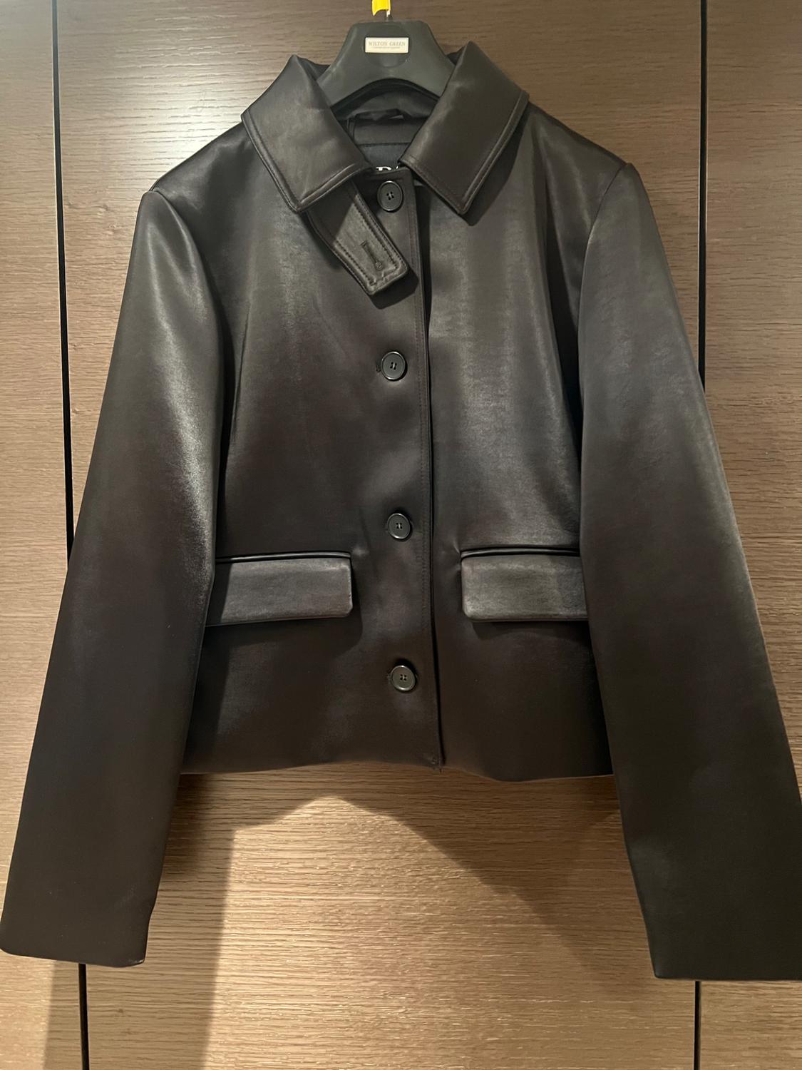Black leather jacket hanging on a black hanger with a wooden background. It has front buttons and two pockets.