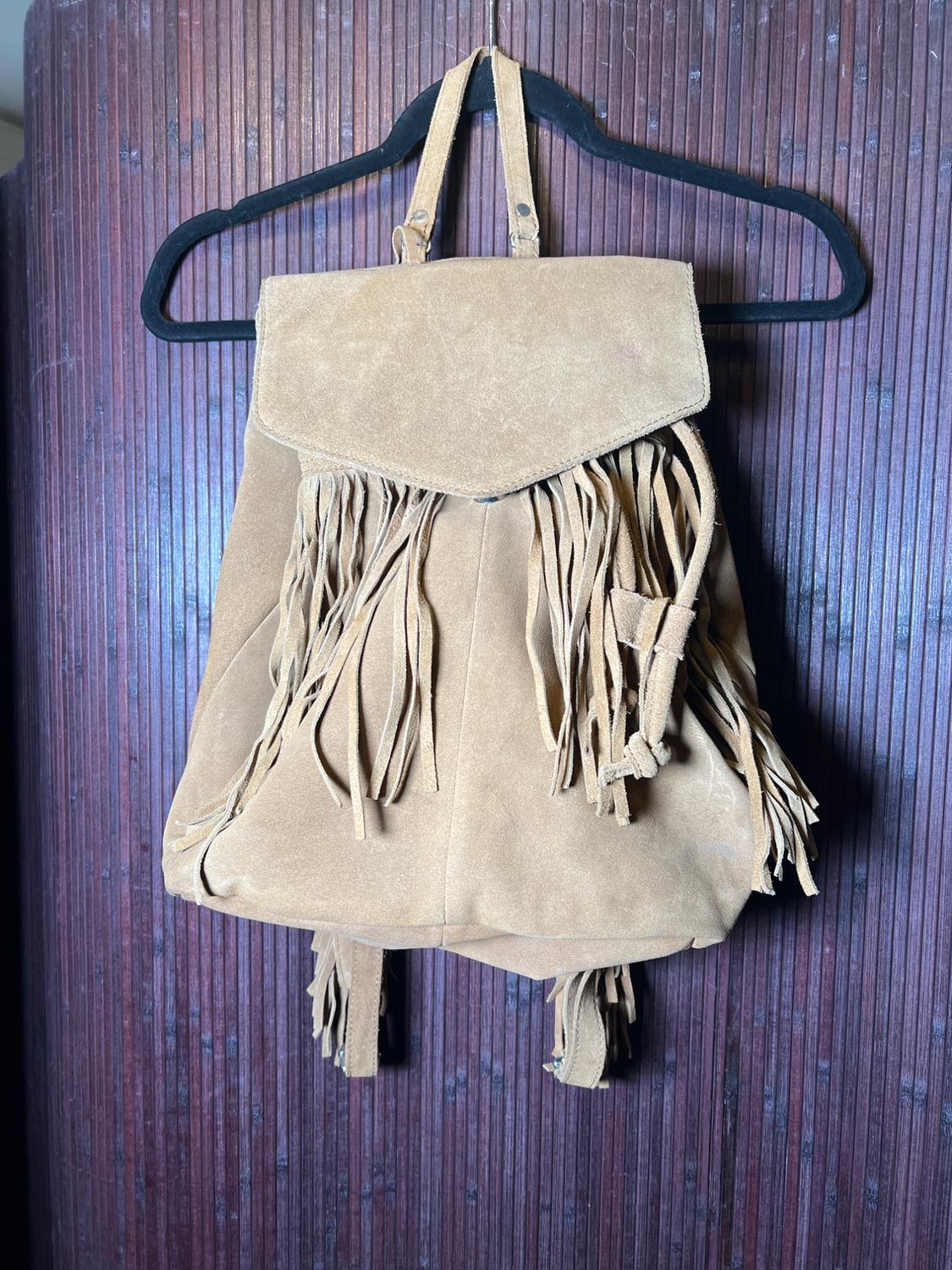 Beige suede backpack with fringe hanging from a black hanger against a dark brown wooden background.