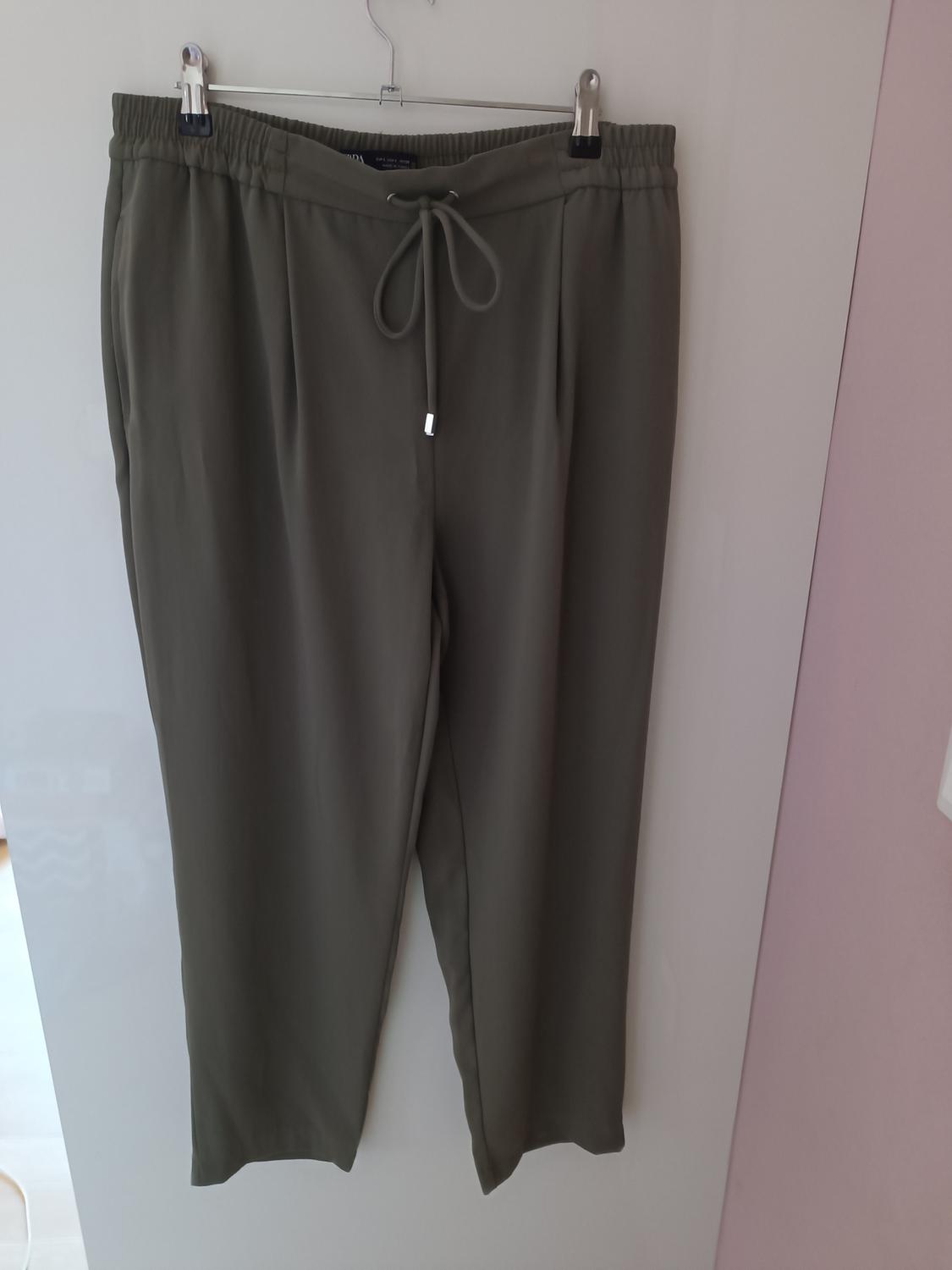 Olive green trousers hanging on a metal hanger, with elasticated waistband and adjustable drawstring, against a white and pink background.