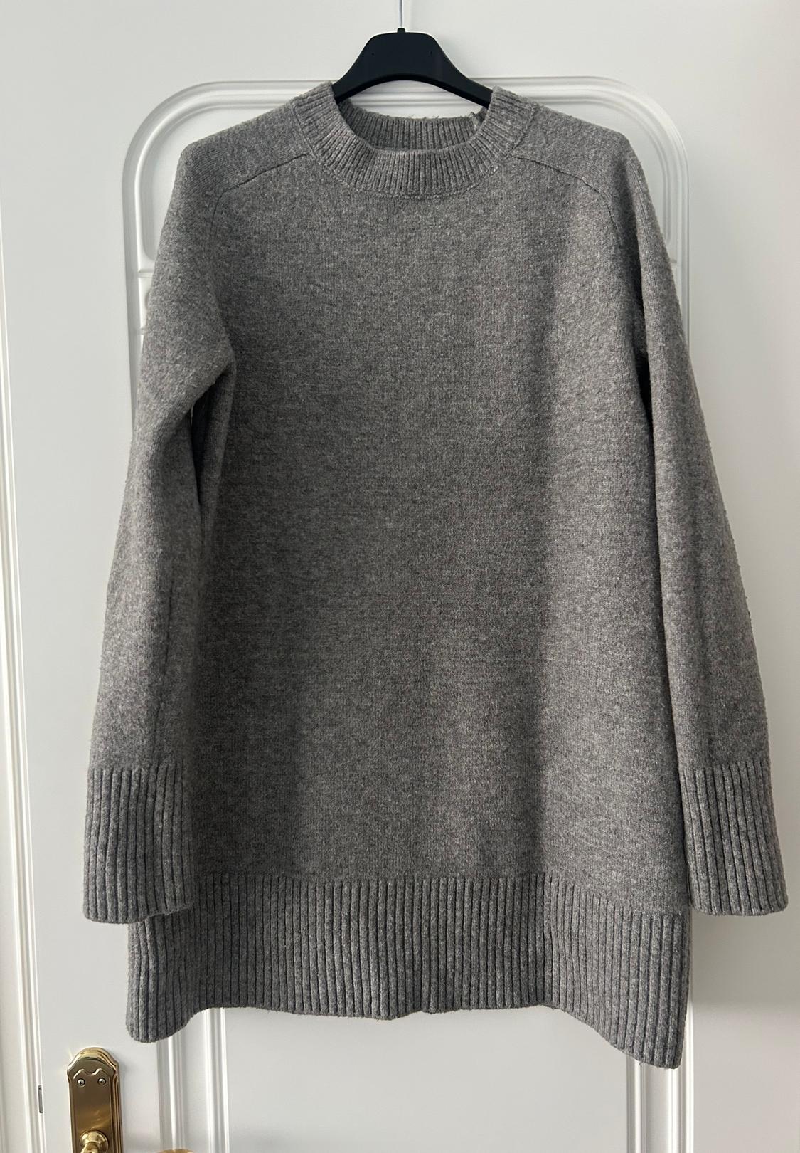 Chunky knit grey jumper hanging on a black hanger over a white door, with ribbed detailing on cuffs and hem.