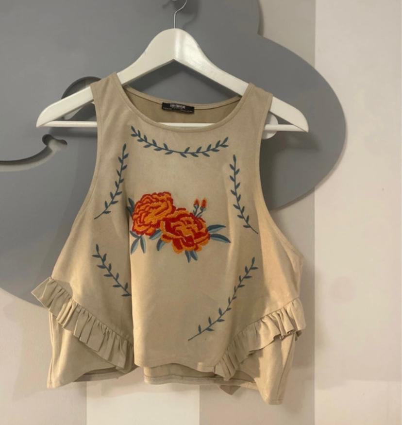 Top beige sleeveless with orange floral embroidery and green leaves, hanging on a white hanger against a grey background.