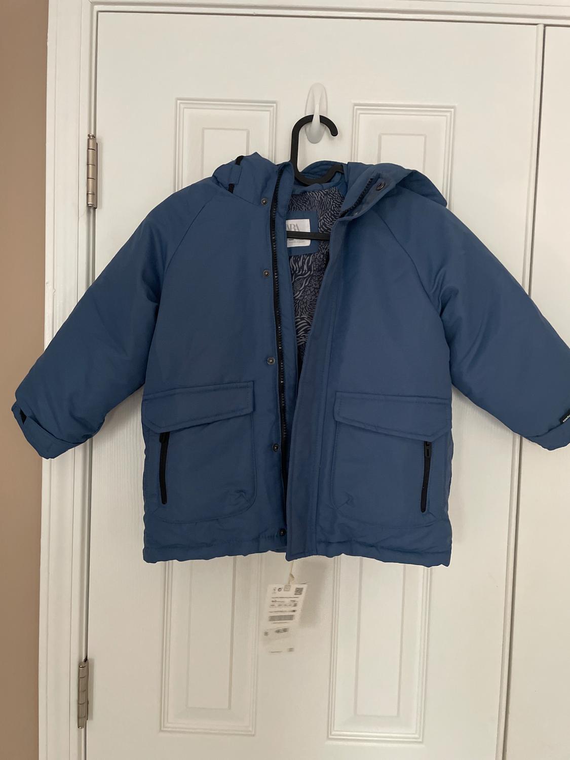 Blue hooded jacket hanging on a white door. It has pockets with black zippers and a visible hang tag.
