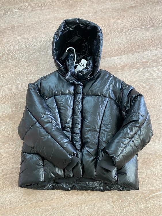Black quilted jacket with hood on light wood floor. Visible tag on collar with plastic hook.
