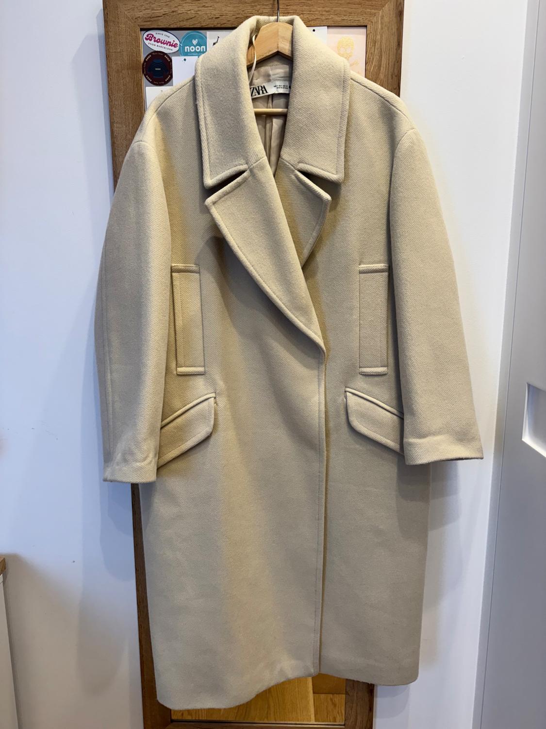 Beige coat hanging on a wooden hanger, with wide lapels and pockets, against a white background with stickers.