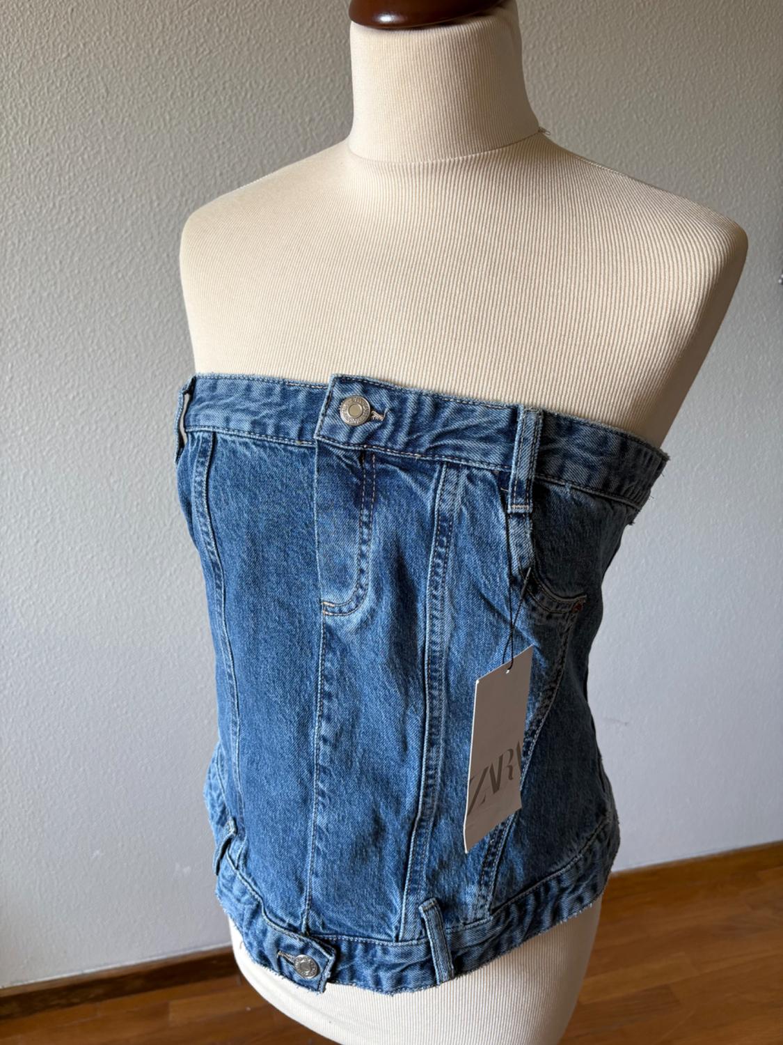 Blue strapless denim top with buttons and belt loops on a mannequin. Label with the text ZARA. Grey background and wooden floor.