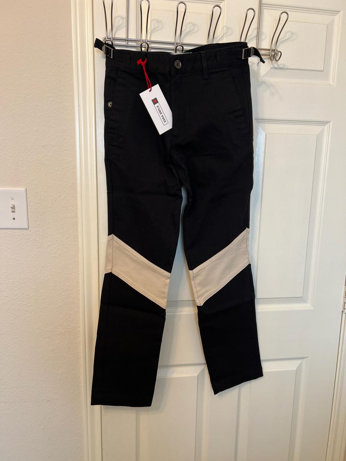 Black trousers with a beige V-shaped stripe hanging from a clothes rail on a white door. Label with the text ZARA SRPLS.
