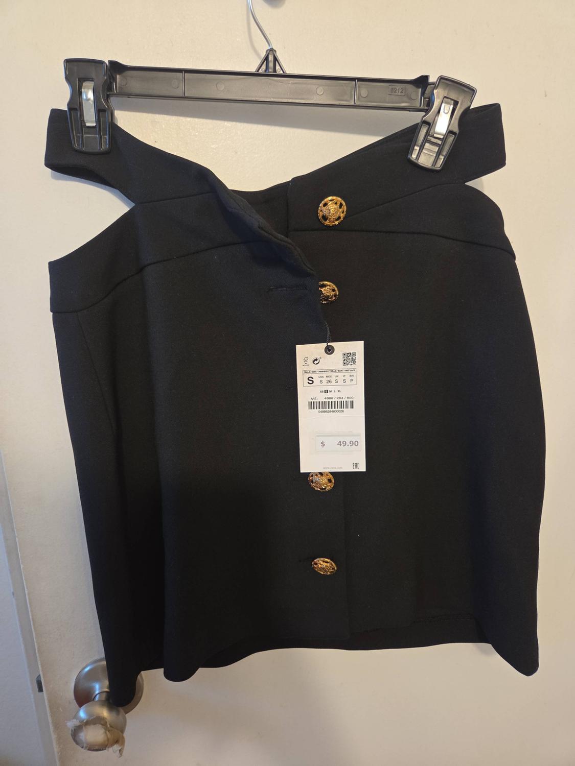 Black skirt with cutouts and gold buttons hanging on a white door. Tag with text: S 26 S S P $49.90.