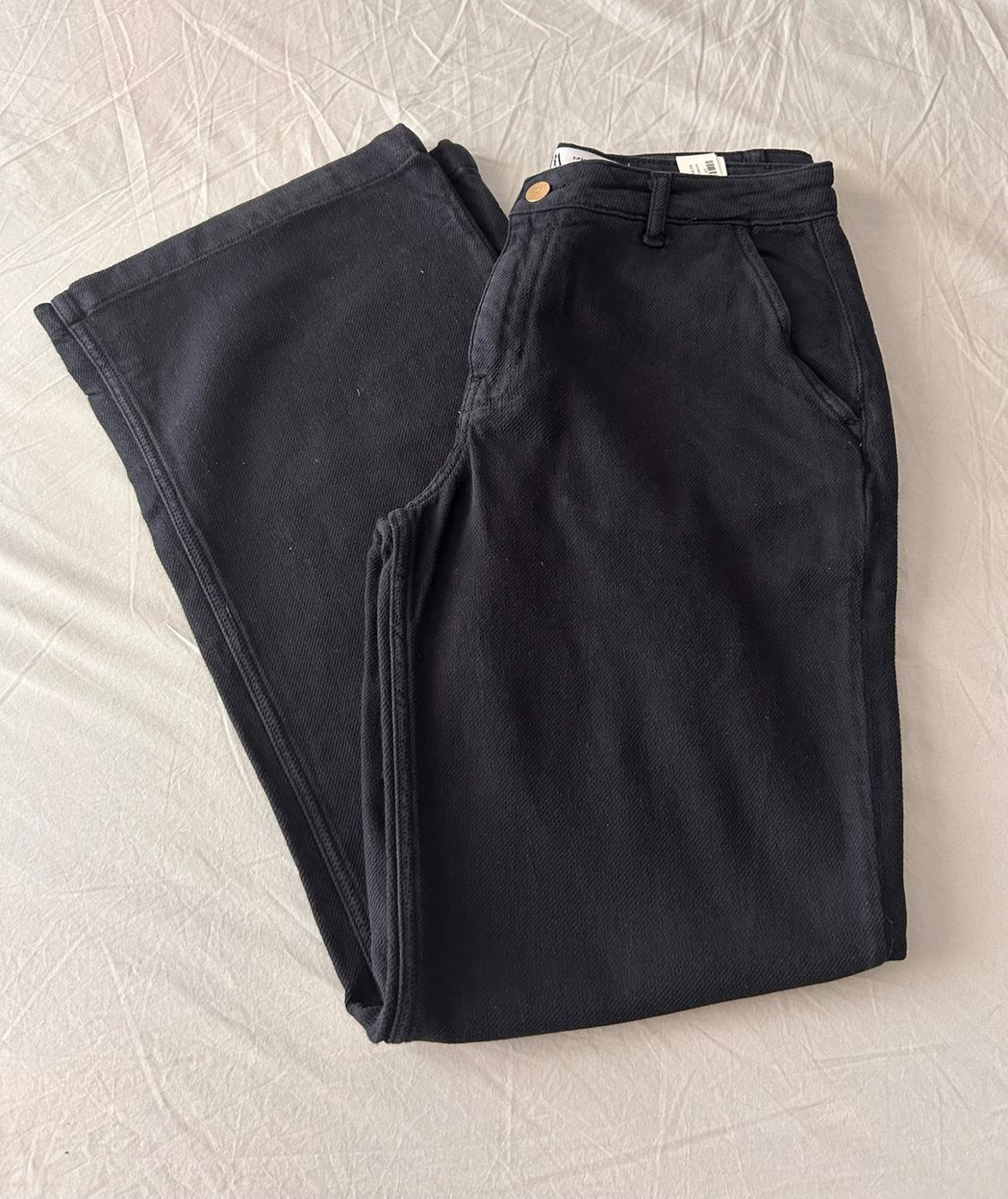 Black trousers folded in half on a crumpled white sheet. The waistband with a metal button, belt loops and pockets is visible.