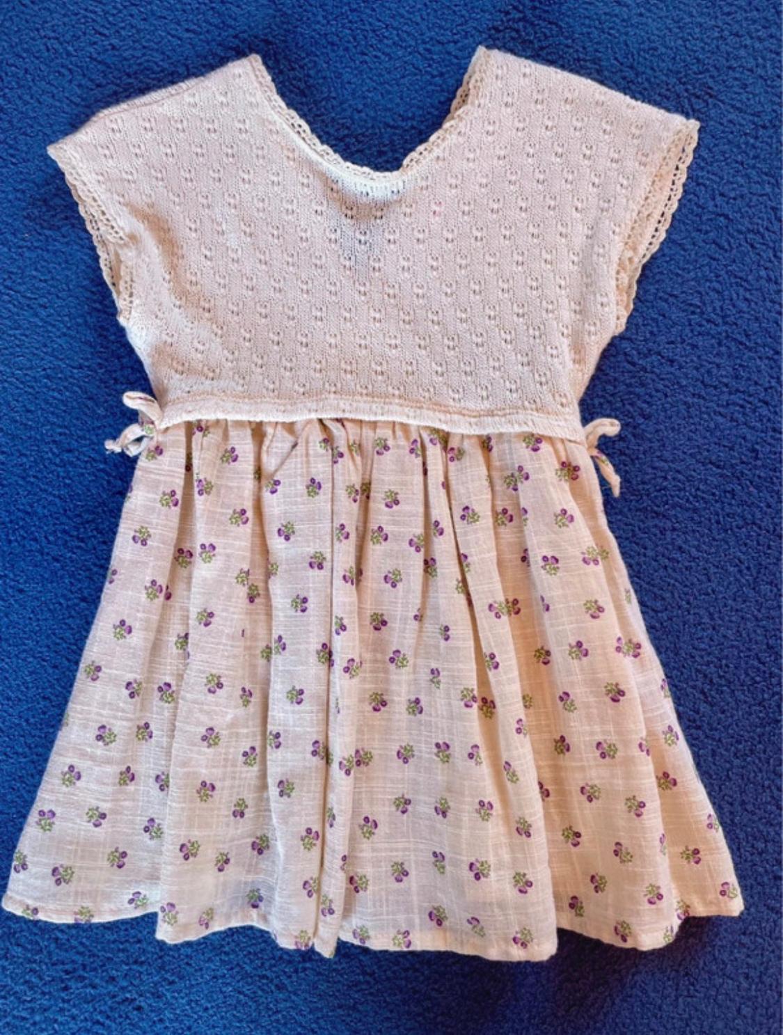 Child's dress on a blue background, with a white lace top and a skirt with a purple and green floral print.