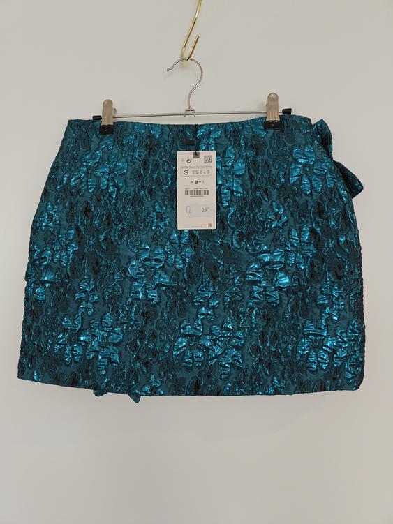Metallic blue crinkled mini skirt, hanging on a hanger with a visible tag showing size S and price €29.