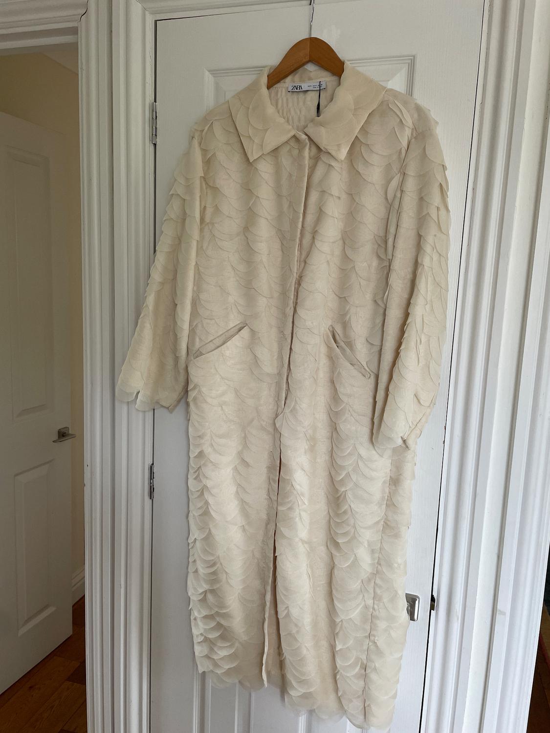 Long beige coat with a scale-like texture, hanging on a wooden hanger over a white door. Visible tag: ZARA.