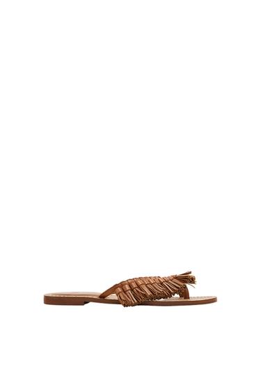 Flat brown sandal with braided design and fringes on the upper, on a minimalist white background.