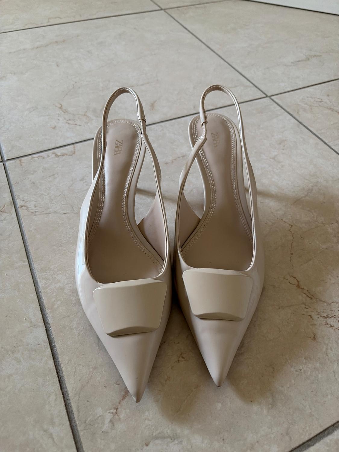 Beige pointed-toe slingback heels with a front embellishment and ZARA text on the insole, on a tiled floor.