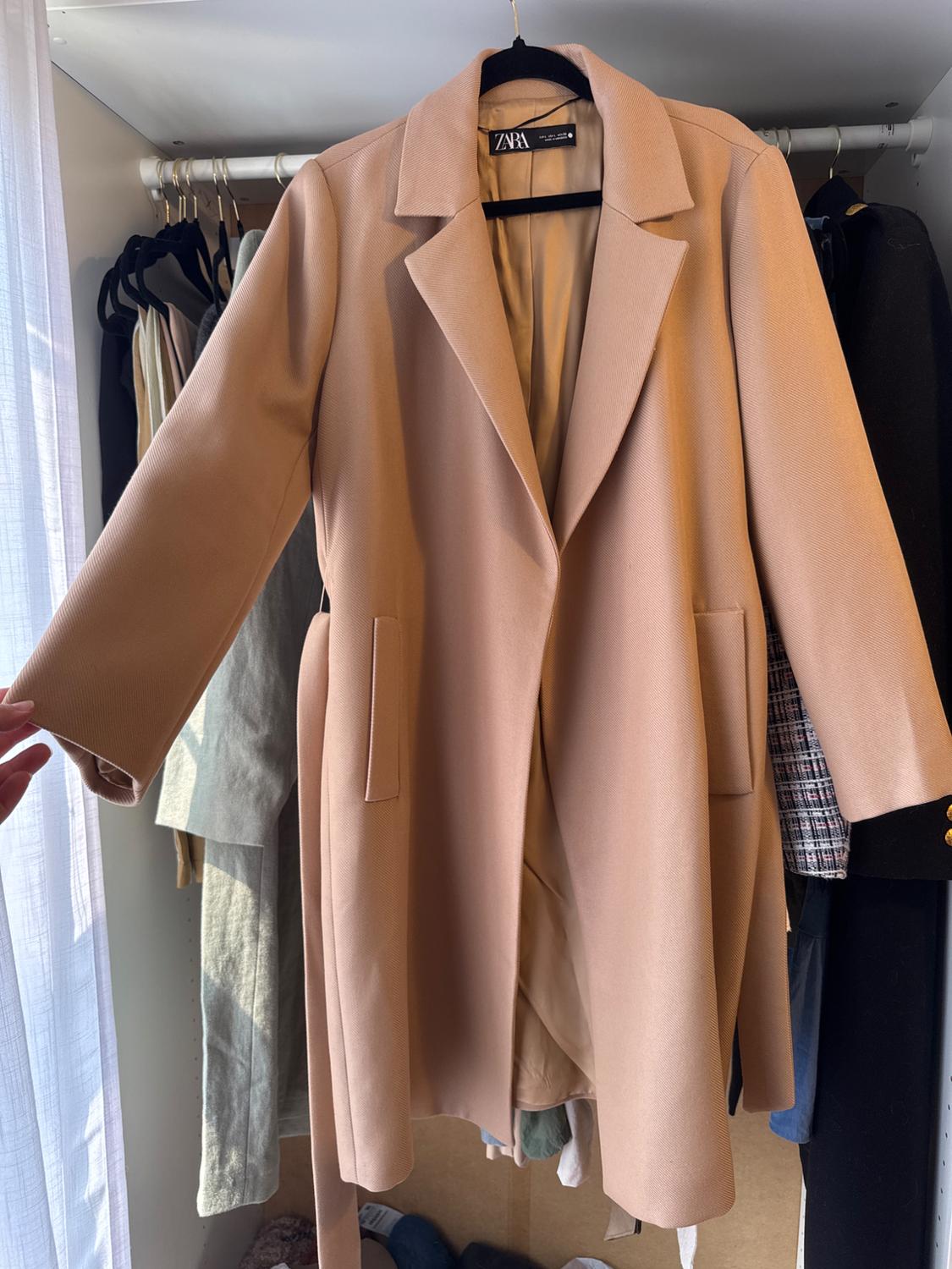Beige long coat with a ZARA label, featuring a lapel collar and pockets, hanging in a wardrobe with natural light. A hand can be seen holding the sleeve.
