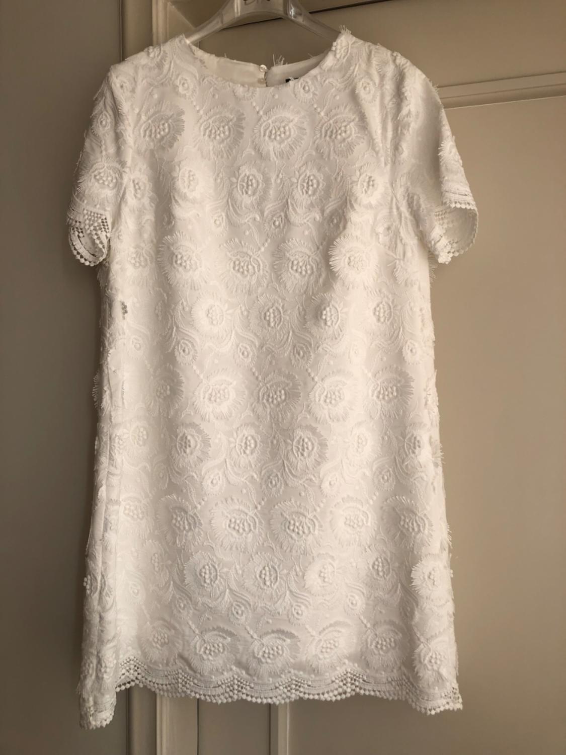 White lace dress with floral embroidery, short sleeves and crochet detailing on the edges, hanging on a hanger in front of a beige door.