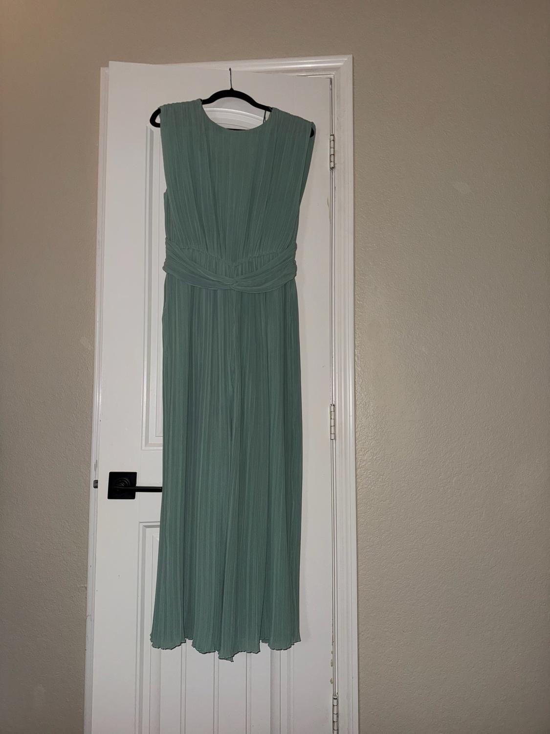 Green sleeveless pleated jumpsuit with waist detailing, hanging on a hanger against a white door. Casual and relaxed style.