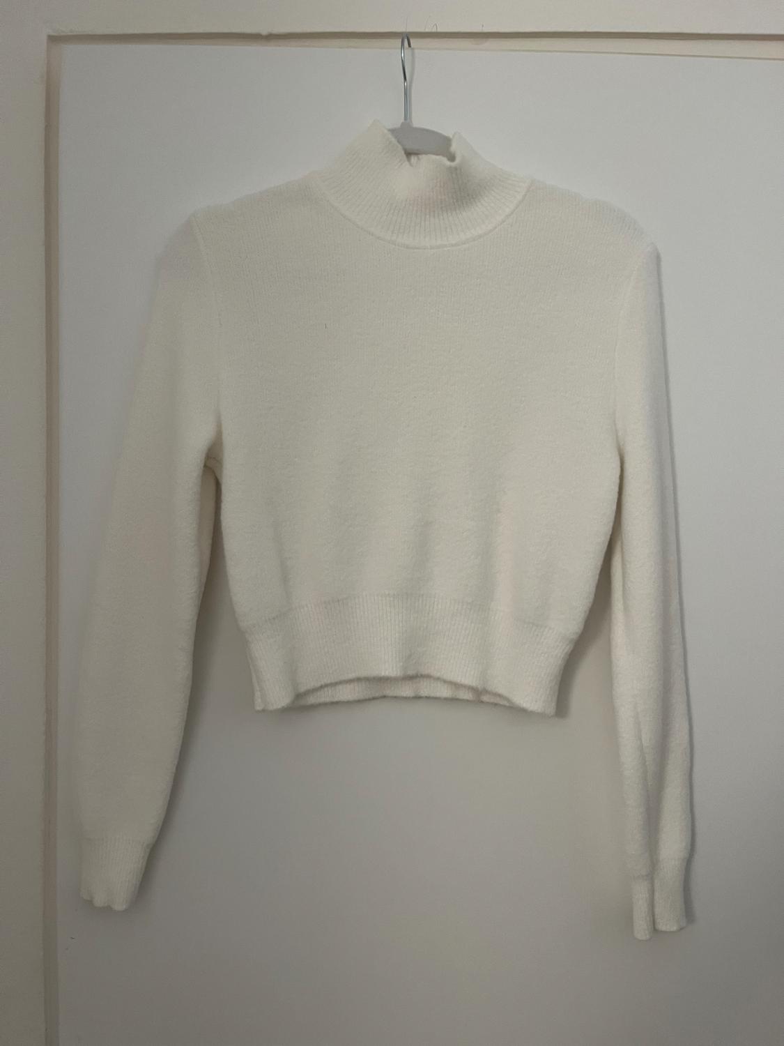 White long-sleeved roll-neck jumper hanging on a hanger against a white wall.