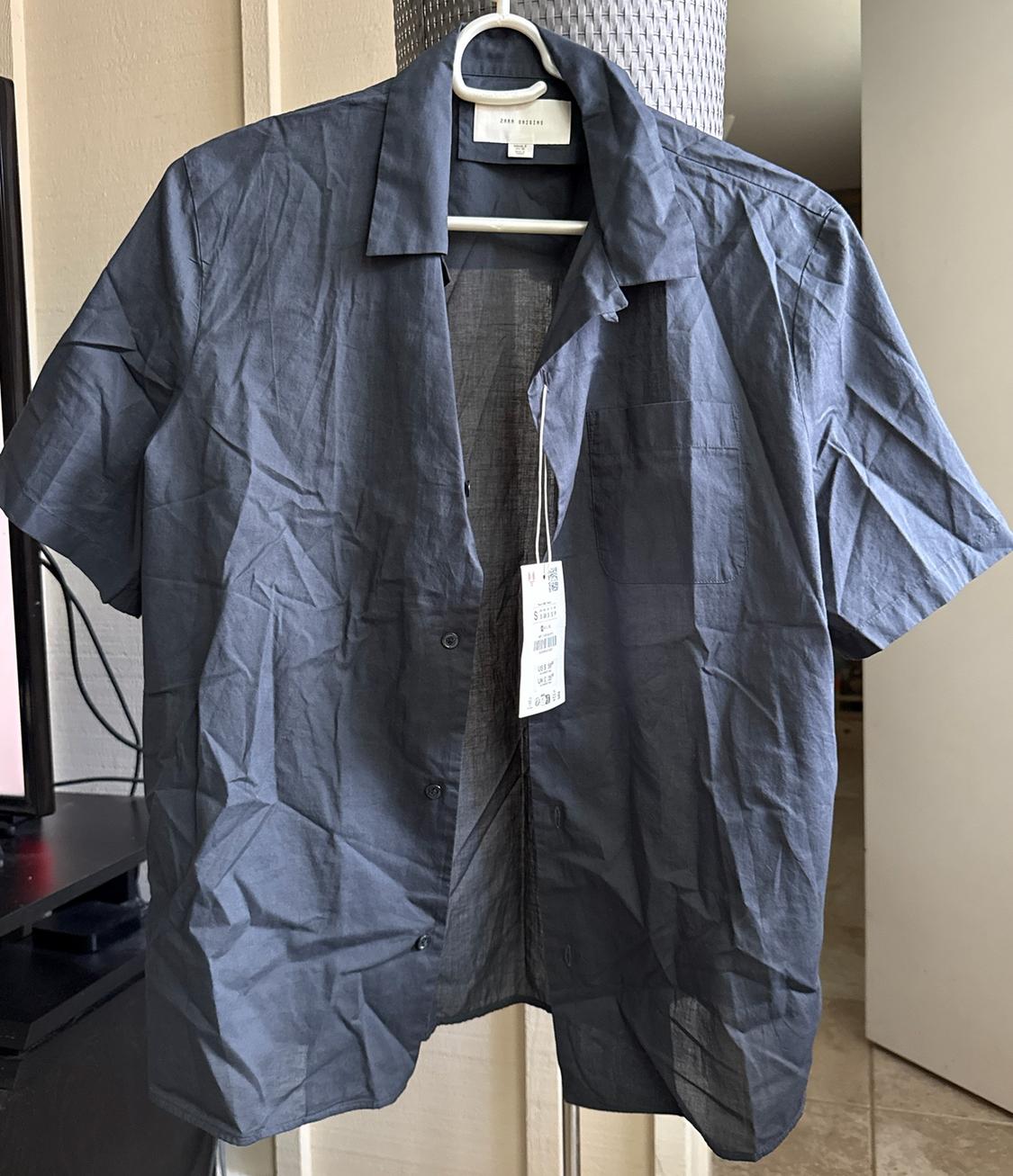 Unbuttoned, wrinkled dark gray short-sleeve shirt on a white hanger. ZARA ORIGINS text on the collar. Indoor setting.