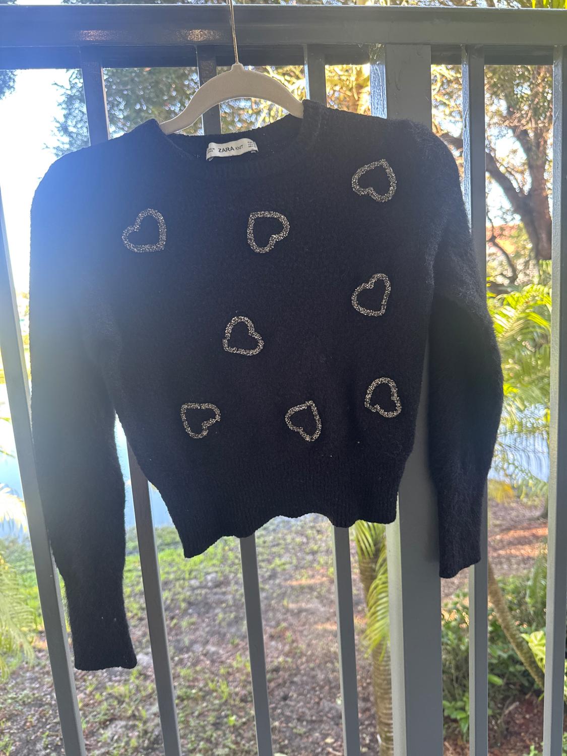 Black sweater with glittery hearts hanging on an outdoor balcony, surrounded by greenery and natural light.