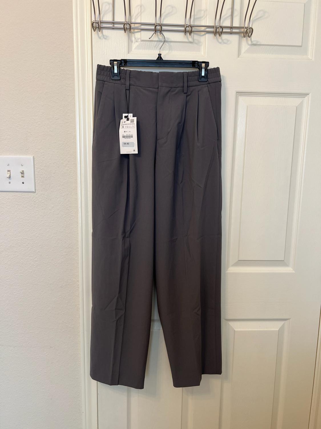 Dark grey pleated trousers hanging on a coat hanger over a white door. They have a label bearing the text S and 39.90.