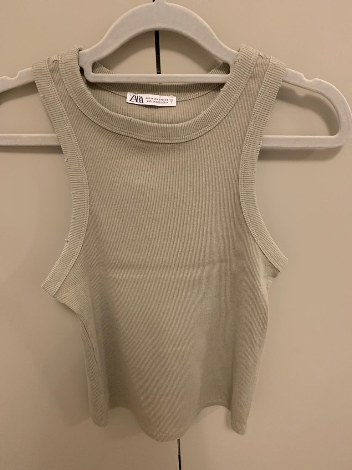 Beige ribbed vest top hanging on a grey hanger. Visible label with the text "Zara". Light beige background.