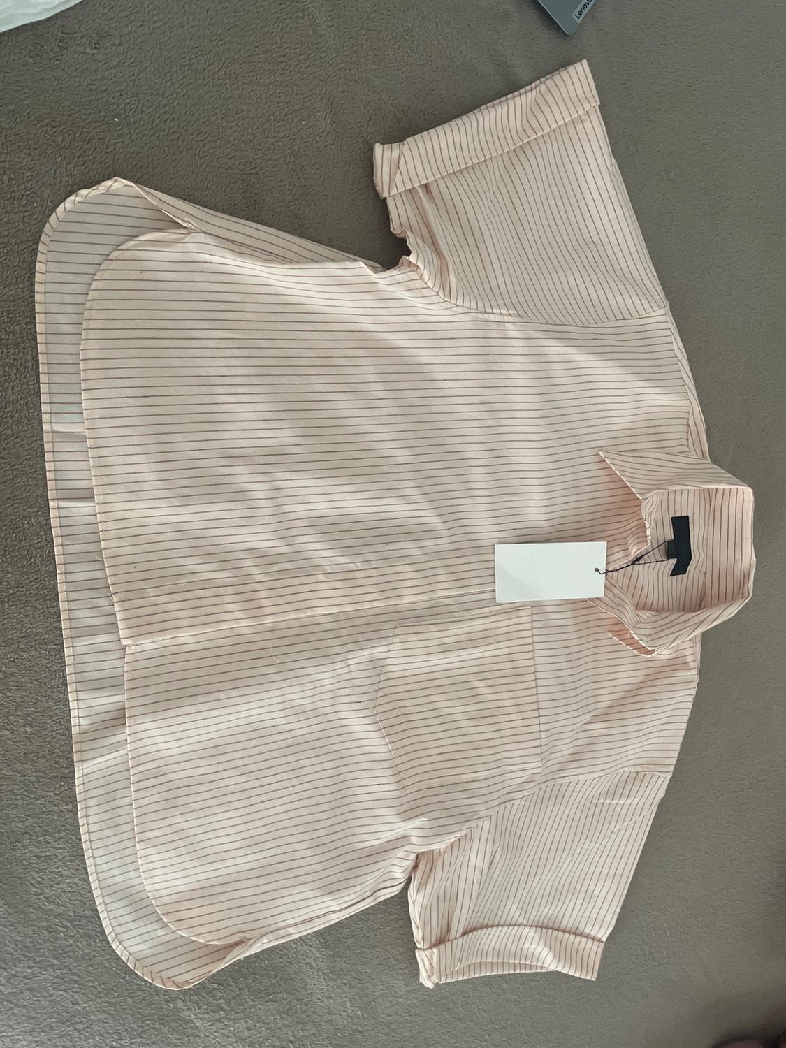 Light pink short-sleeved shirt with fine stripes and a pocket. It is laid out on a grey fabric surface and has a white label.