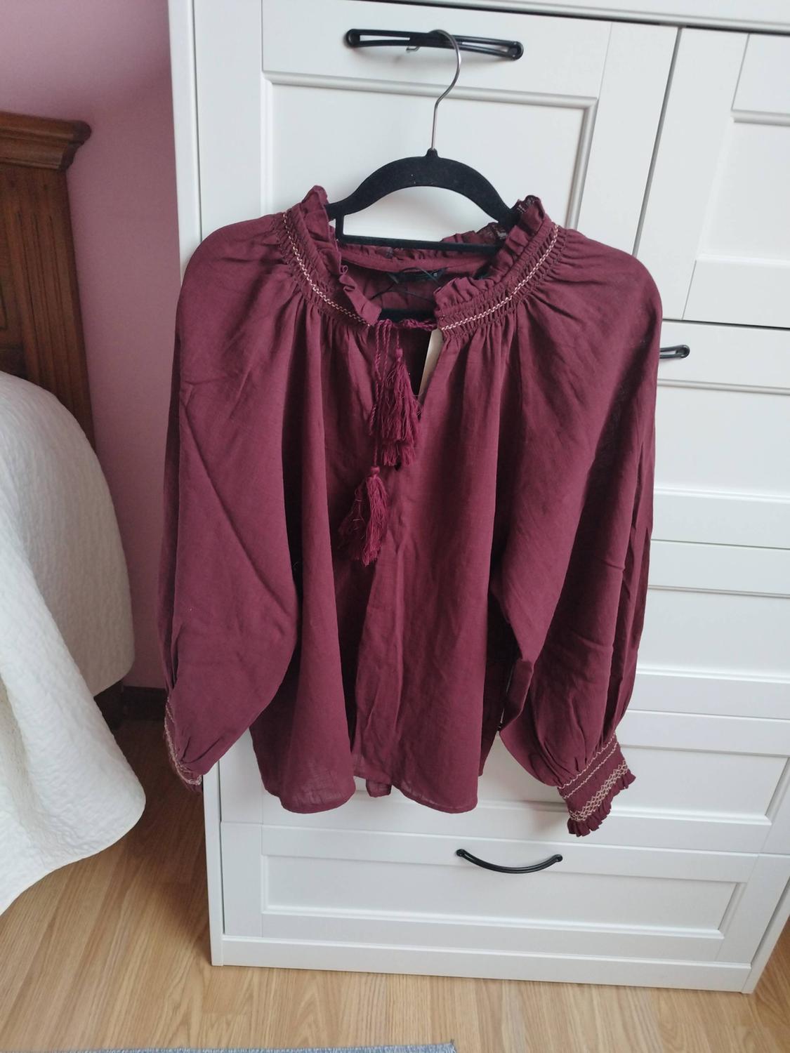 Burgundy long-sleeved blouse with a ruffled collar and tassels, hanging in a white wardrobe in a relaxed bedroom setting.