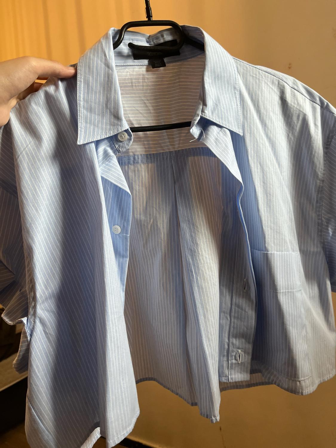 A hand holds an unbuttoned short-sleeved shirt with blue and white stripes on a black hanger. Warm background.