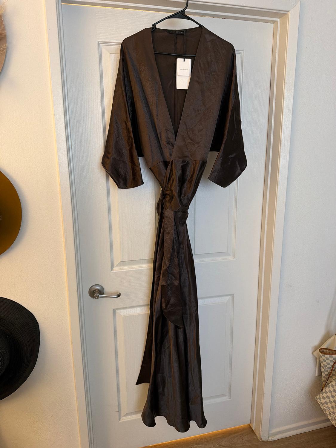 Long brown dress hanging on a white door, with visible tag. Indoor setting with black hat and bag on the sides.
