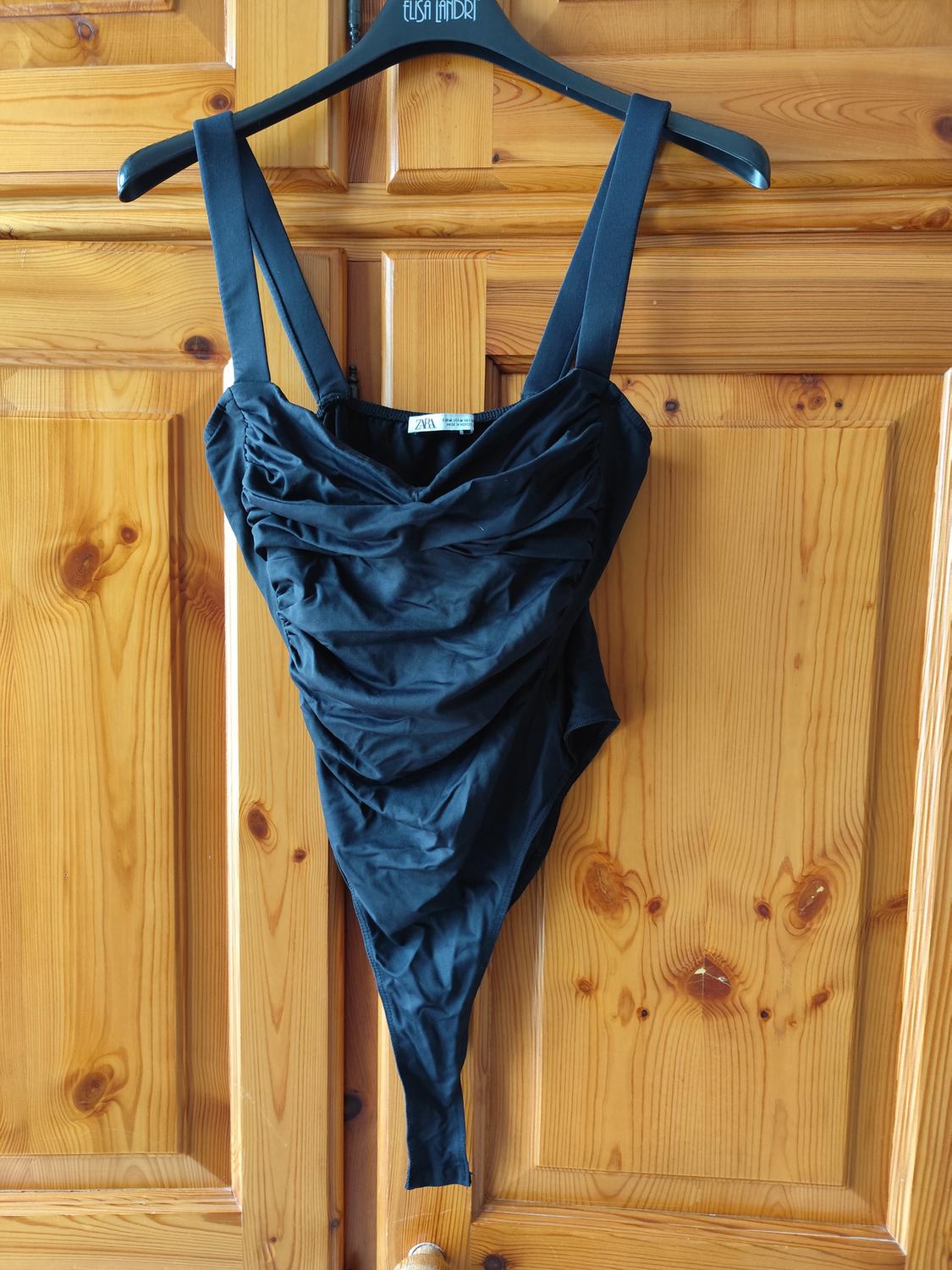 Black bodysuit with wide straps hanging on a black hanger over a wooden door. Visible label: "ZARA".