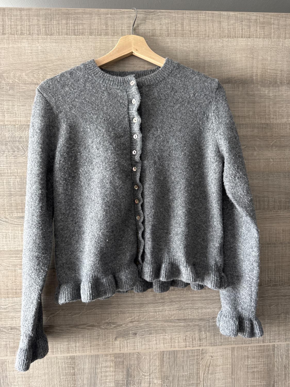 Grey wool cardigan with white buttons, hanging on a wooden hanger against a light wooden background.