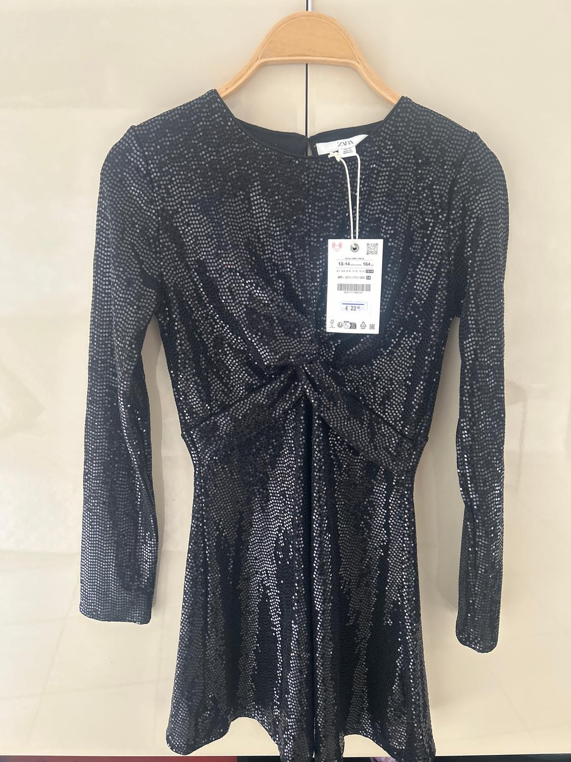Shiny black dress with long sleeves and a knot detail on the front, hanging on a wooden hanger. Visible tag.