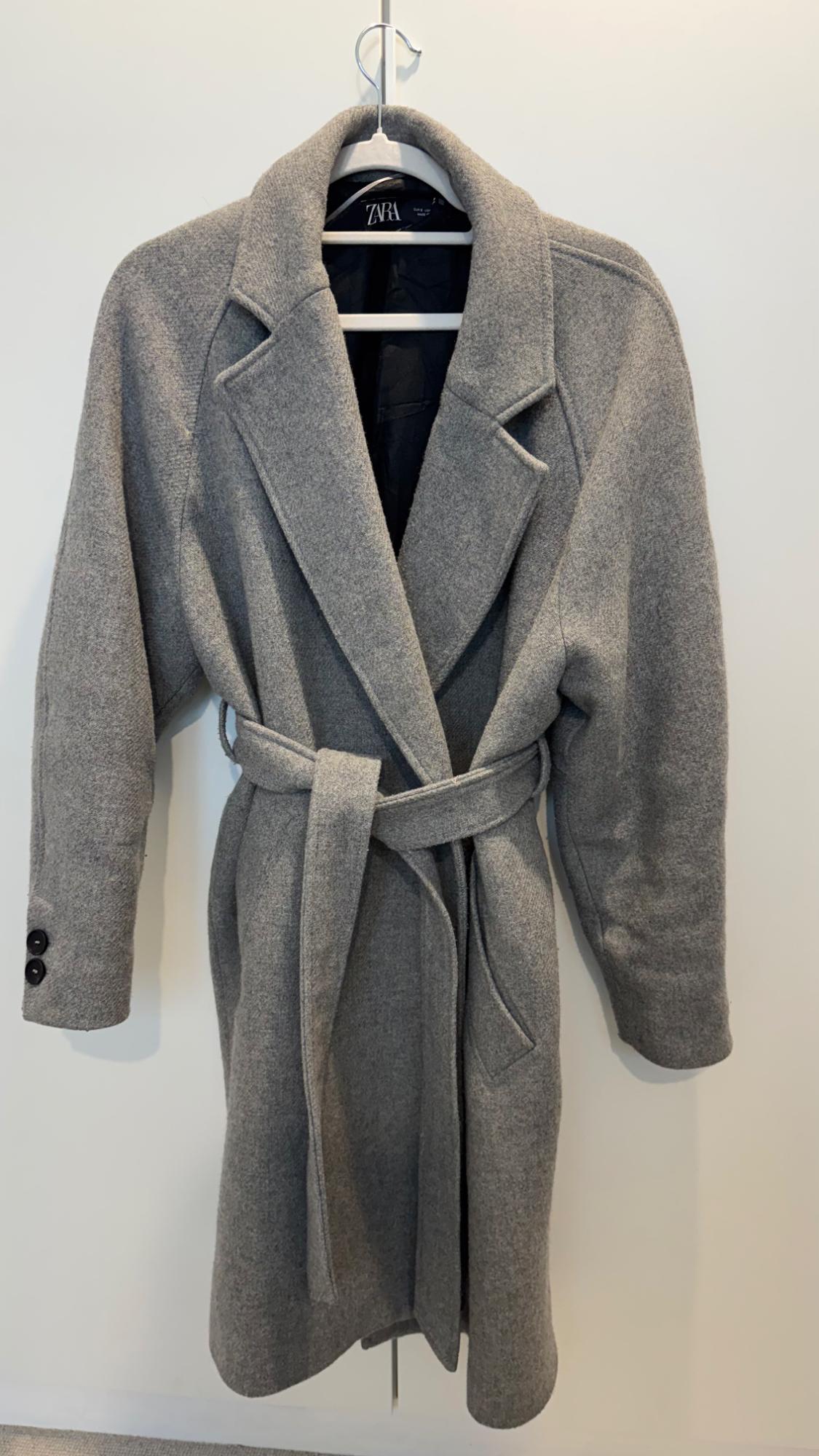 Grey coat with belt hanging on a white hanger. Visible label with the brand "ZARA". White background.