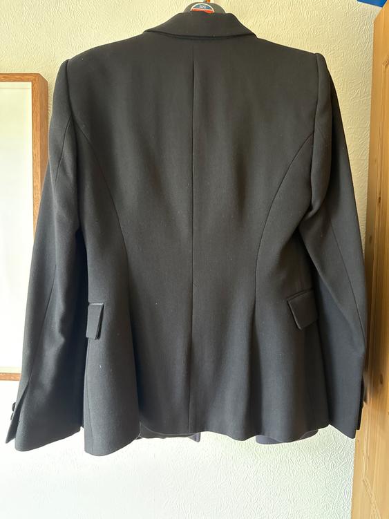 Black jacket hanging on a hanger, viewed from behind. Textured white wall background with a wooden frame to the left.