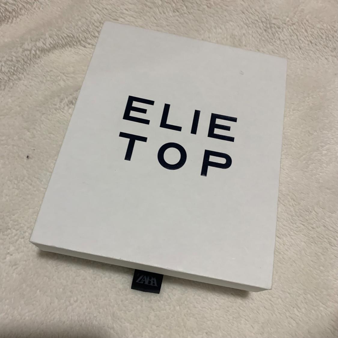 White box with "ELIE TOP" text in black on a beige fabric surface. A black label with "ZARA" protrudes from the box.