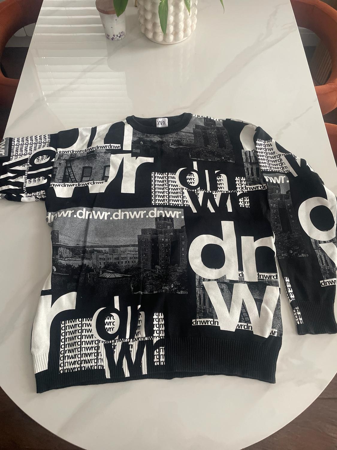 Black sweater on a white table with urban prints and white letters. Texts: ZARA, dnwr.dnwr.dnwr.dnwr., dr, W.