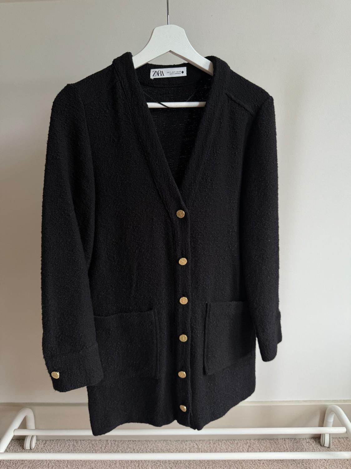 Black knitted jacket with gold buttons, front pockets and visible Zara label, hanging on a white hanger.