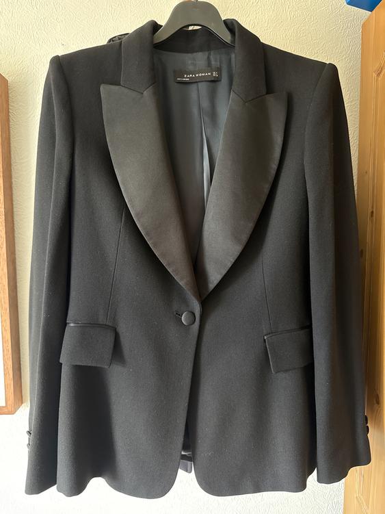 Black women's jacket with satin lapels, hanging on a hanger. Visible label: "ZARA WOMAN". Light wall background.