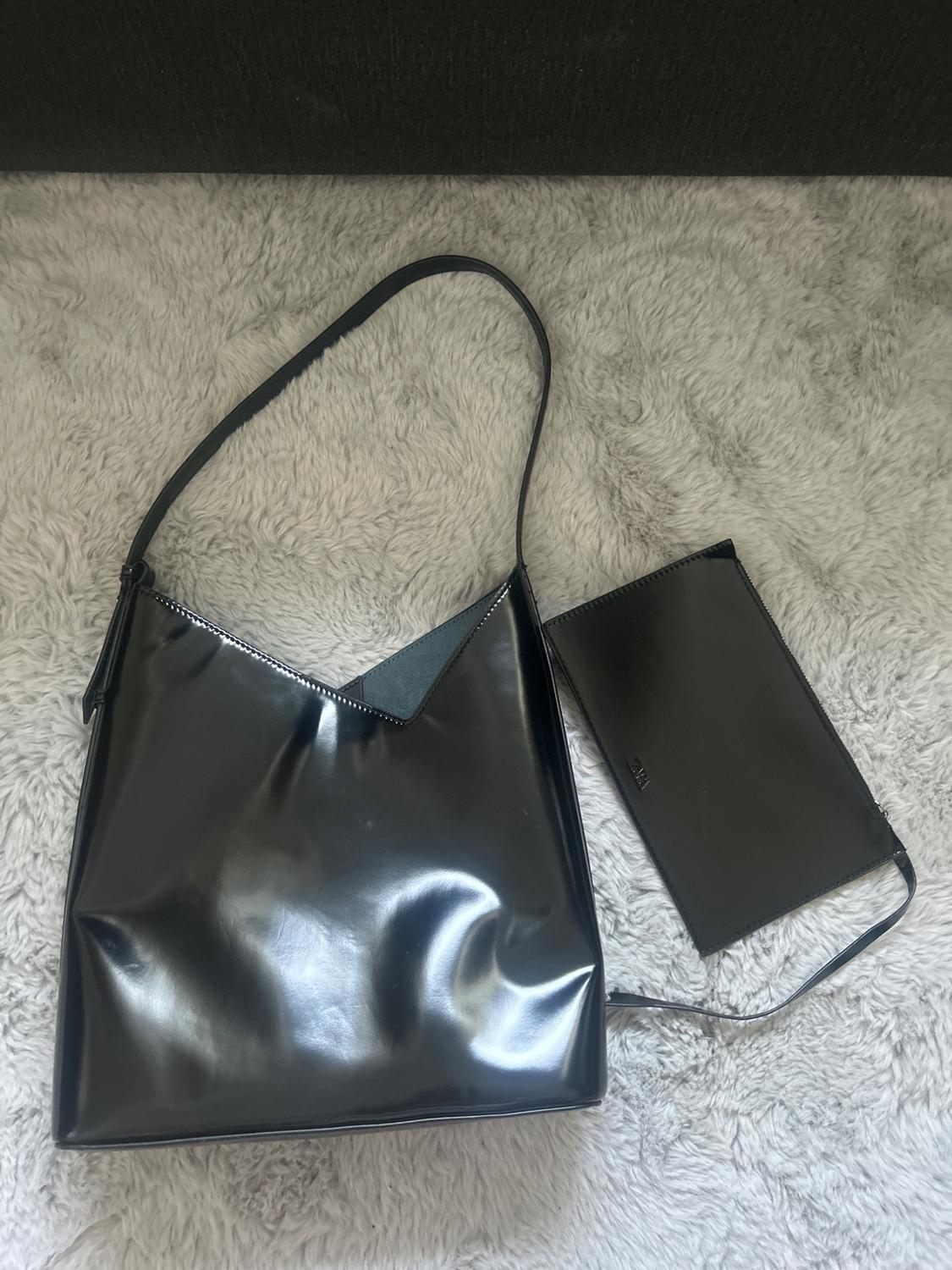 Glossy black bag with a V design and matching purse with the text ZARA, on a light grey shag rug.