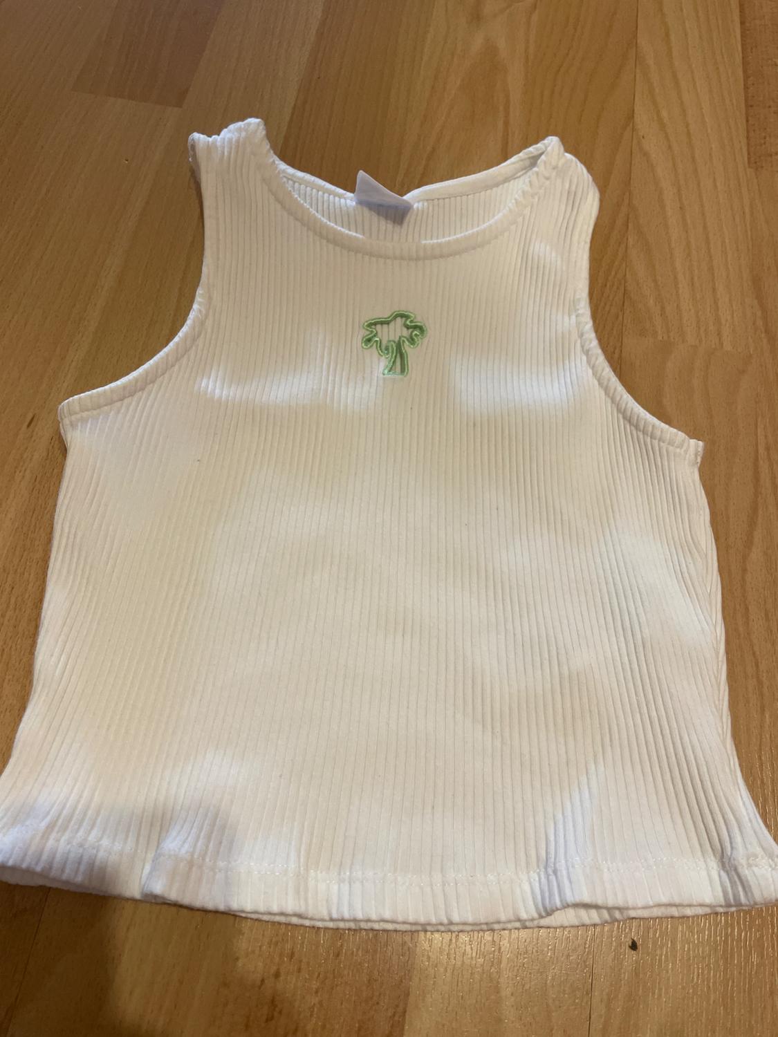 White ribbed vest top with small green palm tree embroidery on the chest, on a light wood background.