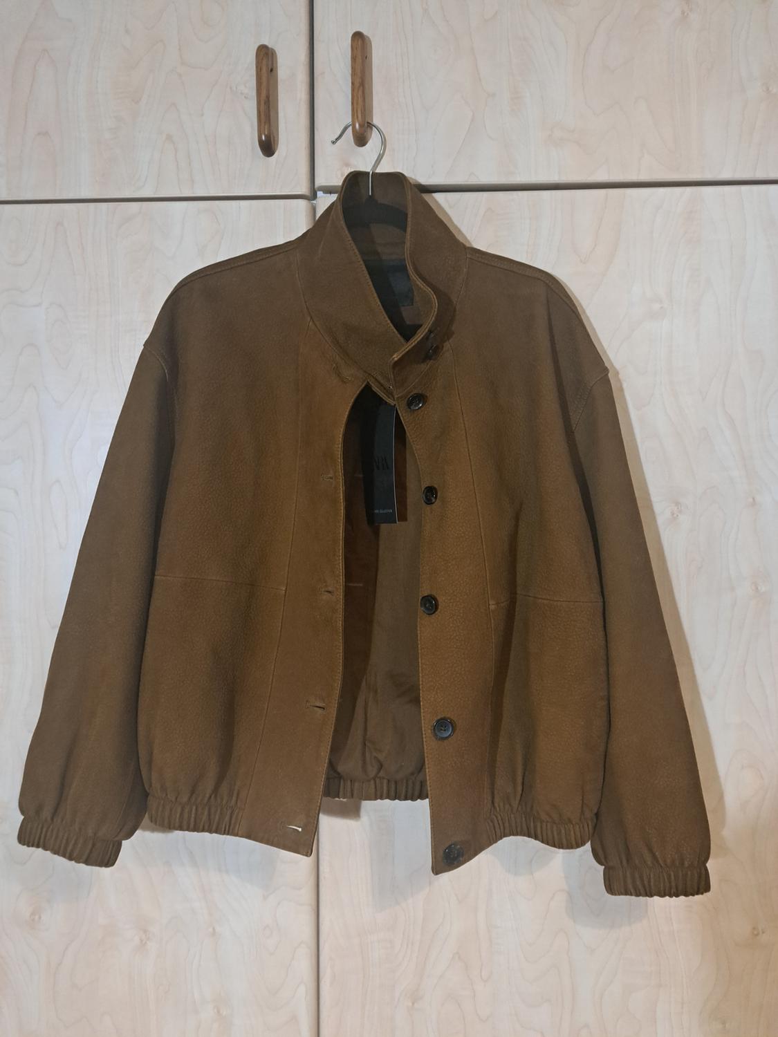 Brown jacket hanging on a wooden hanger, with black buttons and elasticated cuffs, against a light wood wardrobe background.